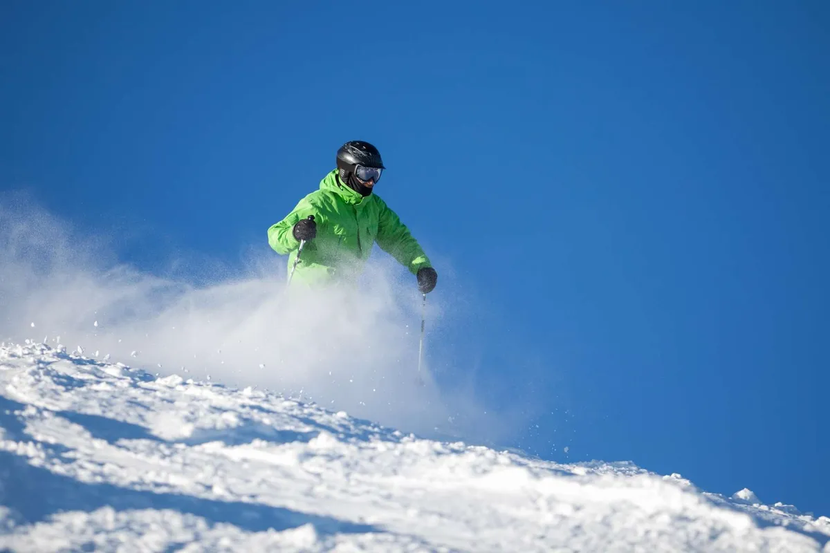 A vibrant winter scene with a skier in a bright green jacket gliding down a sunlit, snow-covered slope against a clear blue sky. The image radiates excitement and accessibility for all levels of winter sports.