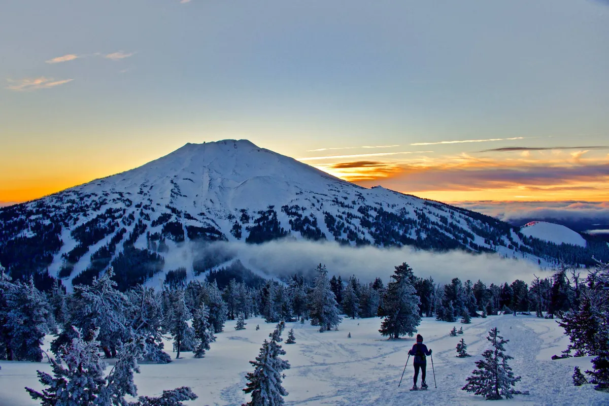 A dramatic winter landscape featuring a snow-covered mountain, a skier making their way down the slope, tree-lined ridges, and a sky glowing with sunset colors. The atmosphere is adventurous and invigorating, capturing the spirit of winter sports and breathtaking natural beauty.