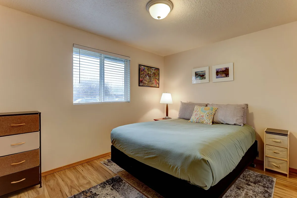 A serene bedroom with a cozy queen bed, nightstand, and lamp, illuminated by sunlight streaming through a window. Artwork above the bed and gentle color tones complete the restful ambiance.