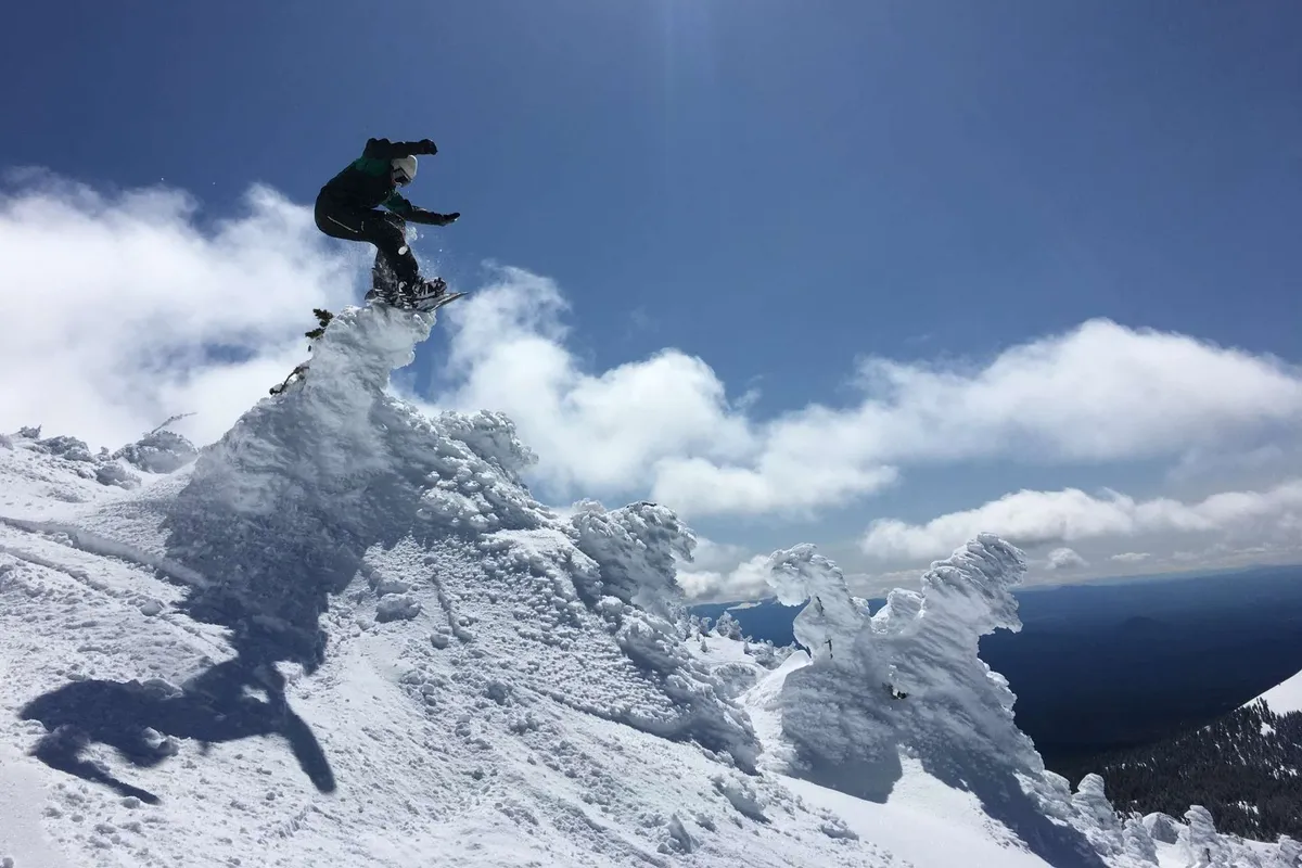 An exhilarating exterior shot of a snowboarder catching air off a snowy peak with clouds swirling around the summit. The scene is dynamic and energetic, highlighting winter sports and the rush of mountain adventure.