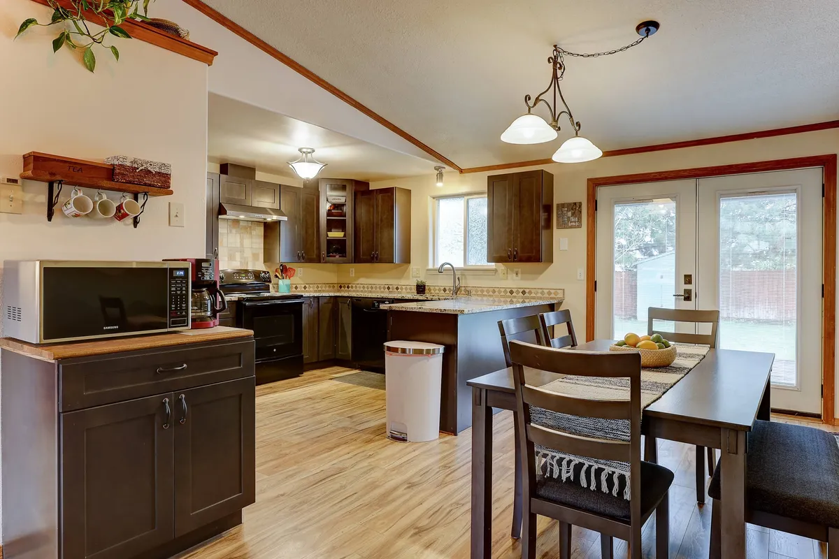 This image features a bright open-plan setting, showcasing a spacious dining table adjacent to a kitchen island with modern cabinets and large windows letting in natural light. The scene invites togetherness, with clear space for family meals, entertaining, and cooking.