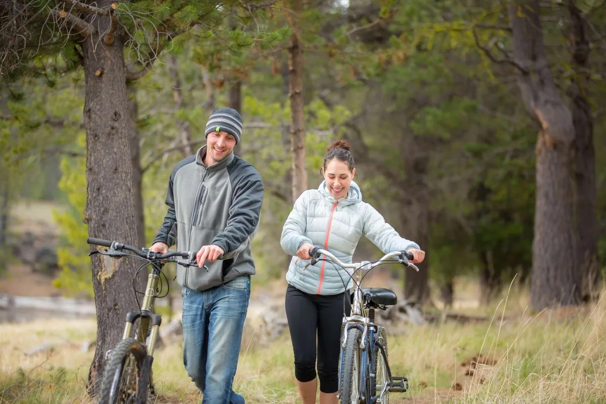 This exterior scene captures a couple walking with two bicycles along a peaceful, tree-lined path. The duo is engaged in conversation, evoking a sense of connection and shared adventure as they explore the outdoors together.