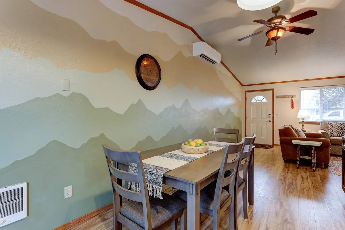 A cozy dining area filled with natural light, featuring a decorative table with fruit, a large mountain mural on the wall, a dining table with chairs, and a wall clock. The setting is perfect for gathering after a busy day or preparing for the next adventure.