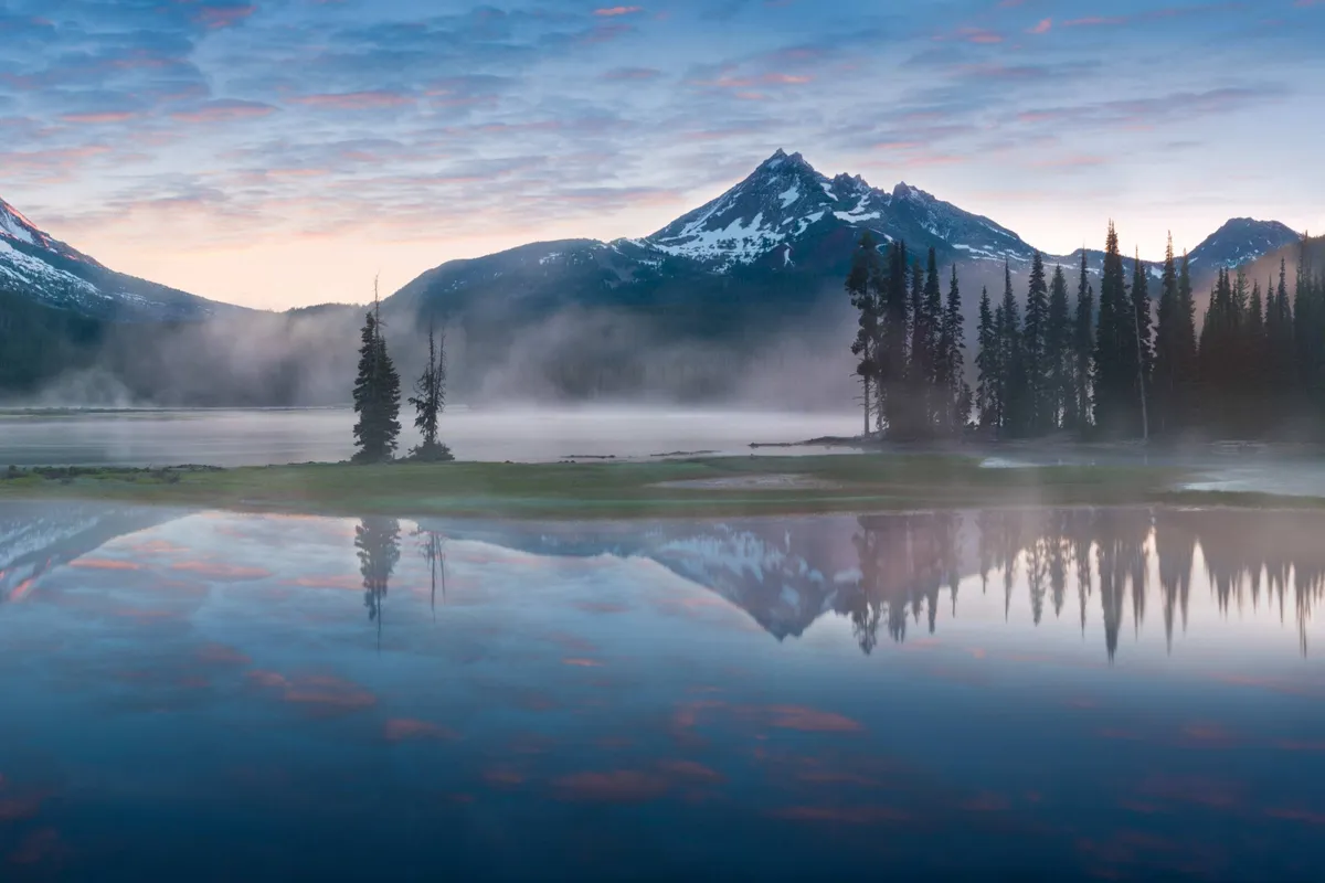 A tranquil lake sits beneath majestic, mist-shrouded mountains and tall evergreen trees, creating a serene and romantic natural backdrop. The stillness of the water and gentle light suggest a peaceful escape, ideal for reflection and connection.