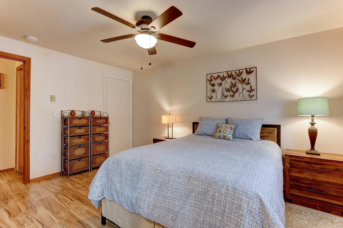 A tranquil bedroom featuring a plush queen bed accented by crisp linens and a decorative wall art above the headboard. A nightstand with a softly glowing lamp and a dresser with additional storage sit nearby, all bathed in natural light, creating an inviting and restful atmosphere.