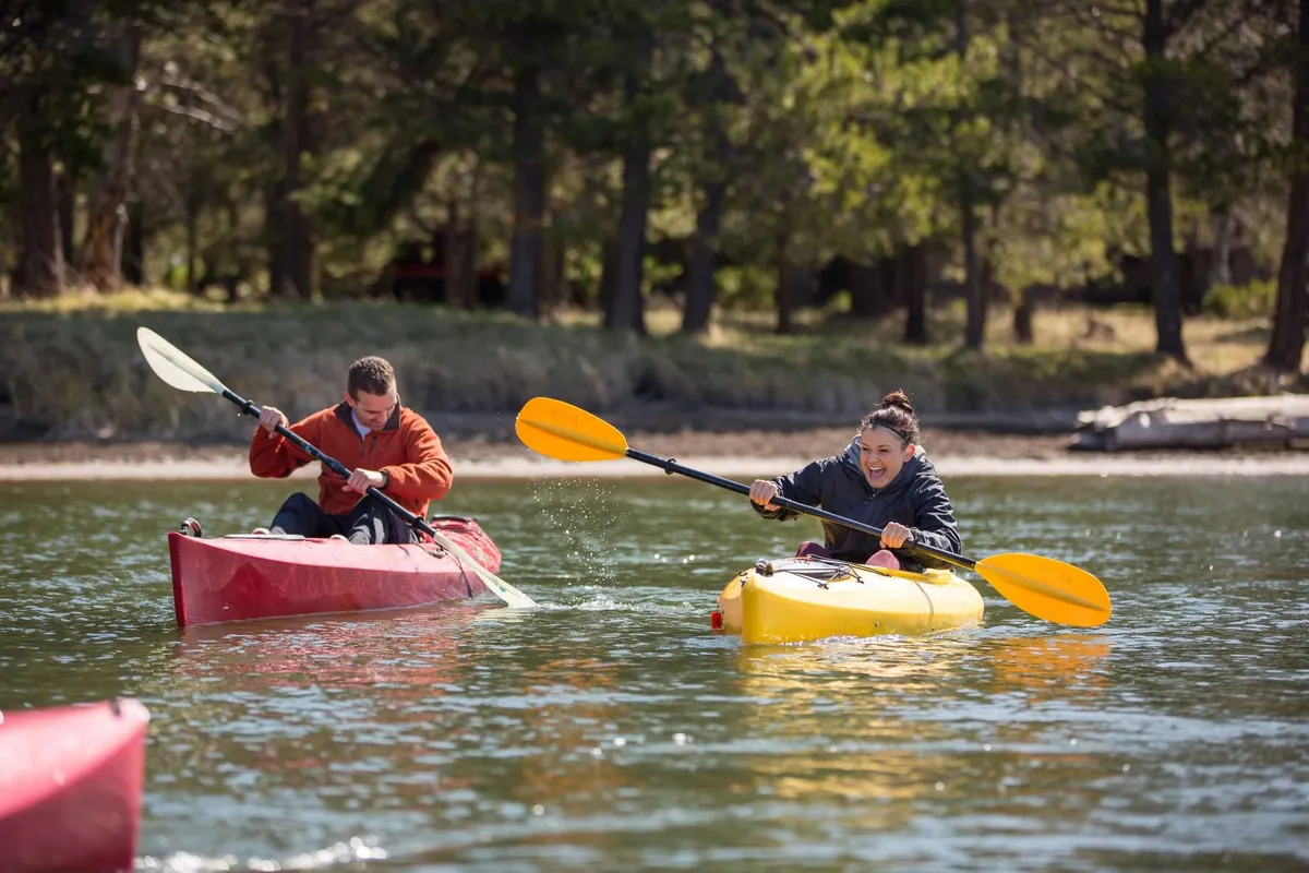 Two kayakers—one in a vibrant yellow kayak, the other in red—paddle across calm water, surrounded by lush greenery and trees. The mood is lively, and the sense of shared adventure is palpable.