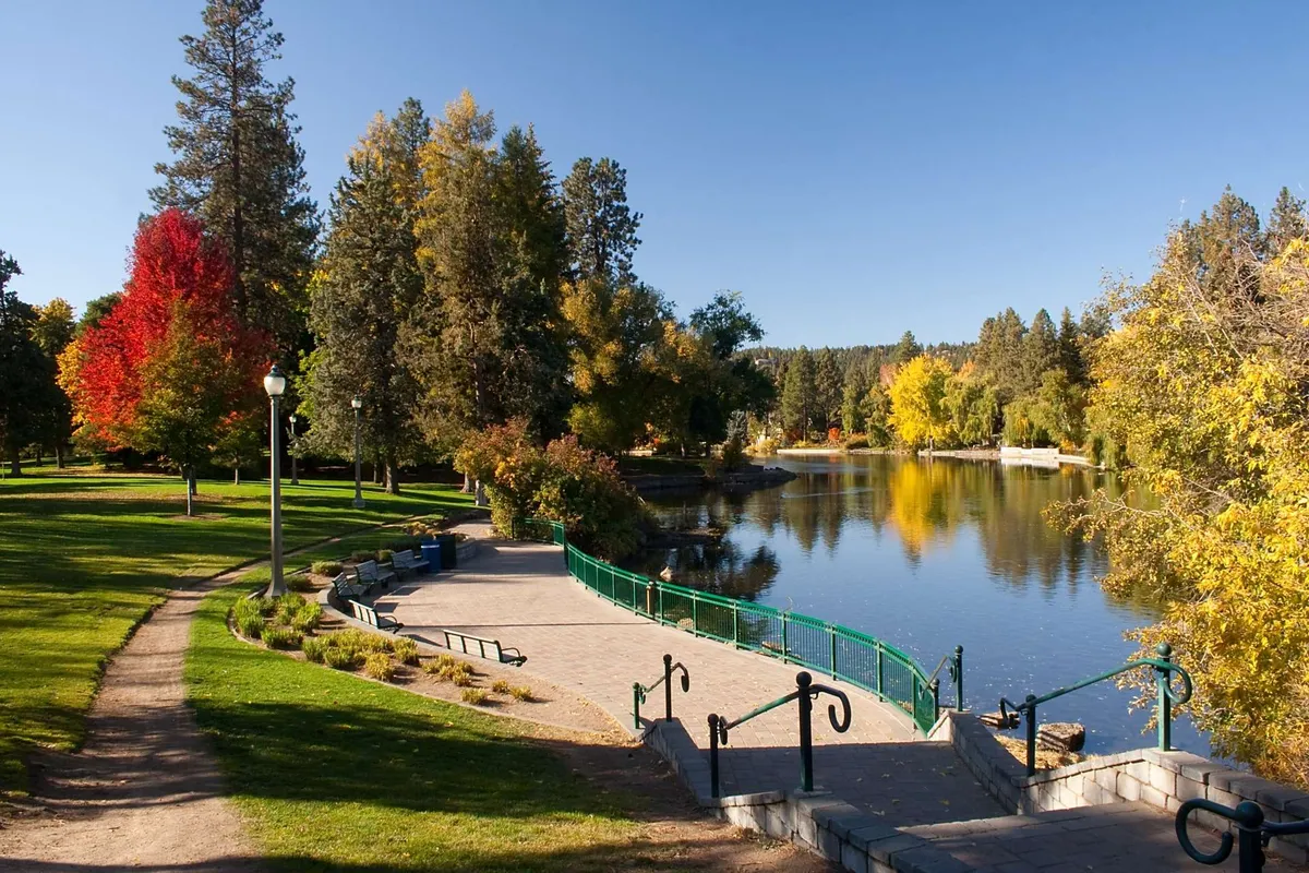 A serene park-like setting featuring a still lake, colorful autumn trees, a winding pathway, and park benches. The environment is peaceful and conducive to outdoor exploration or family picnics.
