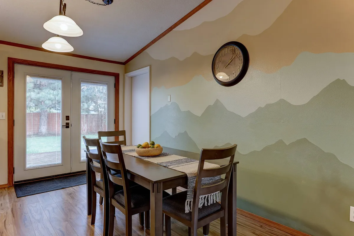A dining area highlighted by a welcoming table, a vibrant mountain mural, and an easy-to-read wall clock. Natural light filters into the space, creating an environment well-suited for both casual meals and focused work sessions.